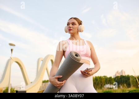 Una giovane donna sicura di sé tiene il suo tappetino da yoga, pronto per una sessione all'aperto. Foto Stock
