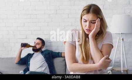 Donna infelice preme la mano per guancia, l'uomo guarda la TV e beve birra dalla bottiglia a casa Foto Stock