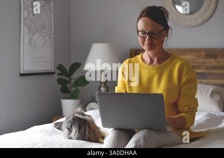 Donna in maglione giallo seduta sul letto con gatto grigio e che utilizza un computer portatile Foto Stock