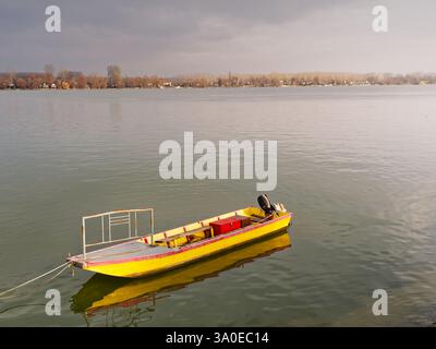 Una barca di colore giallo brillante galleggia sul tranquillo fiume Danubio, con alberi autunnali che costeggiano la costa lontana. Zemun, Belgrado Foto Stock