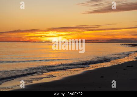Un tramonto mozzafiato dipinge il cielo con vivaci sfumature di arancione e rosa, mentre le dolci onde si infrangono su una spiaggia sabbiosa. La tranquilla atmosfera invita al relax Foto Stock