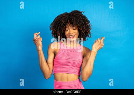 Giovane donna allegra con le dita incrociate che esprime un desiderio a occhi chiusi e espressione di speranza, isolata su uno sfondo blu vibrante Foto Stock