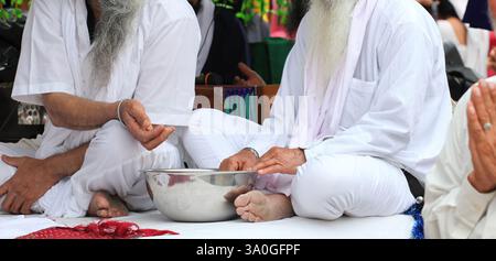 Due capi religiosi Sikh anziani, vestiti con abiti tradizionali bianchi, si siedono a gambe incrociate, eseguendo un rituale con una ciotola di metallo durante un calcio spirituale Foto Stock