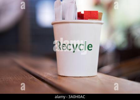 Uno scatto ravvicinato mostra un bicchiere di carta bianca con la parola "riciclo" stampata in verde sul lato, seduta su un tavolo di legno. Foto Stock