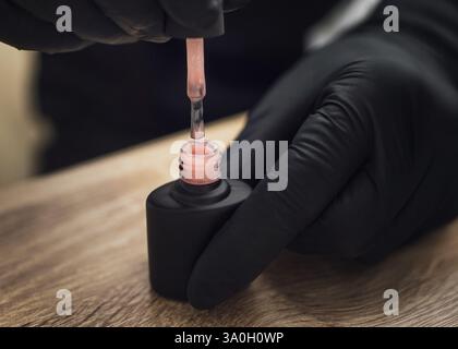 Il tecnico delle unghie che indossa i guanti neri regge un tappo con una spazzola da una bottiglia nera di smalto rosa. Foto Stock