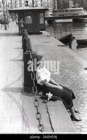 Giovane donna 1954. È seduta al sole a occhi chiusi e gode del bel tempo. Indossa una camicia bianca e una gonna in tessuto Corduroy. Il tessuto a coste, un tipo di cotone con una caratteristica superficie a coste, era popolare negli anni '1950 per la sua durata e calore, rendendolo adatto per l'uso quotidiano come le gonne. Le gonne all'epoca erano spesso lunghe al ginocchio o più, con una linea A o pieghe, che riflettevano lo stile femminile ed elegante degli anni '1950 influenzato da designer come Christian Dior e il suo "New Look" del 1947. Nel 1953, la Svezia era in un periodo di econom post-bellico Foto Stock