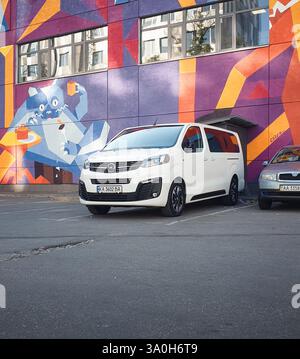 Bianco Opel Vivaro di fronte a pareti colorate. Vista anteriore di tre quarti del furgone Opel Zafira Life in un ambiente urbano, parcheggiato vicino alla berlina compatta. Foto Stock