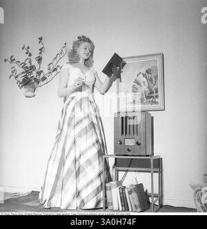 Moda del 1946. Una donna che indossa la tipica moda del dopoguerra. Indossa un abito lungo, il cui tessuto è un motivo di fasce larghe e strette di colori diversi. E' in piedi in una stanza vicino a una radio. Kristoffersson rif. Y127-6 Foto Stock