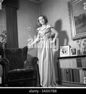 Moda del 1946. Una donna che indossa la tipica moda del dopoguerra. Indossa un abito lungo, il cui tessuto è un motivo di campi stretti e ampi di colori diversi. È in piedi in una stanza accanto a una poltrona. Kristoffersson rif. Y129-3 Foto Stock