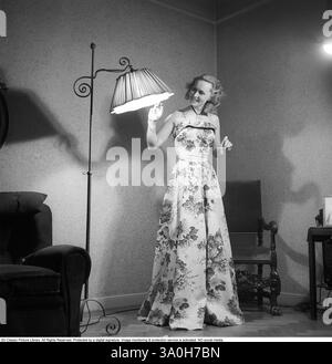 Moda del 1946. Una donna che indossa la tipica moda del dopoguerra. Indossa un abito lungo, il cui tessuto è un motivo di fiori e foglie. È in piedi in una stanza accanto a una lampada da terra che è accesa. Kristoffersson rif. Y129-2 Foto Stock