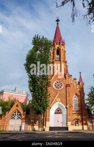 Una suggestiva chiesa in mattoni rossi in stile gotico con alte guglie, una finestra circolare in vetro colorato e una grande porta d'ingresso in legno, circondata dal verde Foto Stock