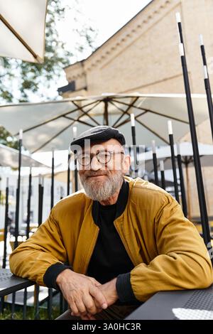 Dapper senior man con una barba elegante e bicchieri rotondi si rilassa all'aperto in un caffè alla moda. Foto Stock