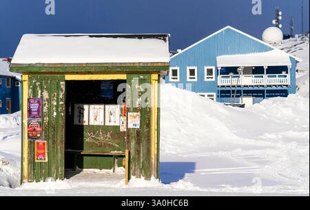 Una colorata fermata dell'autobus riparata dalla neve, con un edificio blu sullo sfondo. La scena cattura un paesaggio invernale con luce solare intensa illumi Foto Stock