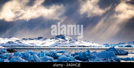 Uno splendido paesaggio di terreno ghiacciato caratterizzato da iceberg galleggianti in un sereno corpo d'acqua, con spettacolari nuvole e raggi di luce solare che si infrangono Foto Stock