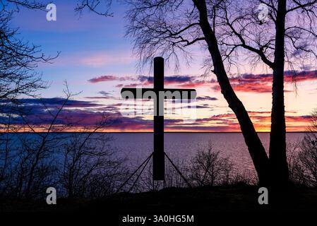 Una grande croce si staglia davanti a un tramonto mozzafiato su un calmo specchio d'acqua in Svezia. Gli alberi incorniciano la scena, esaltando l'atmosfera serena Foto Stock