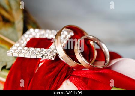 Due fedi nuziali e un favore nuziale gioiello poggiano su un nastro di velluto rosso legato a un bouquet nuziale Foto Stock