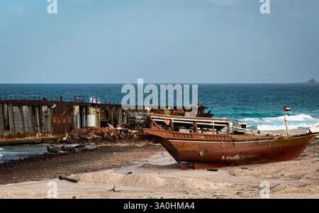 Una barca arrugginita poggia sulla spiaggia sabbiosa di Socotra, con le onde che si infrangono nelle vicinanze e un cielo azzurro limpido sopra la testa, creando una tranquilla vista costiera. Foto Stock