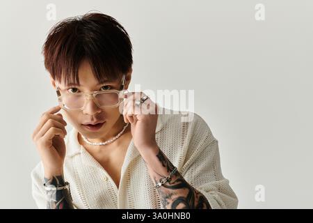 Un giovane e affascinante uomo asiatico regola i suoi occhiali, mostrando un look alla moda e intricati tatuaggi. Foto Stock