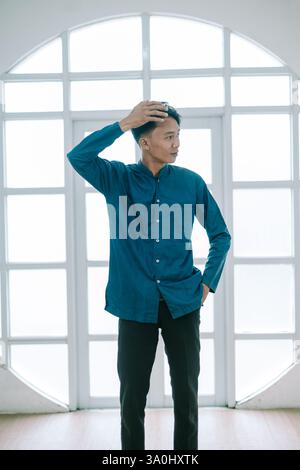 Un giovane uomo con un elegante abbigliamento casual si fa notare in una posa premurosa. Scattata contro una finestra luminosa, l'immagine trasmette un senso di eleganza moderna. Sta in piedi Foto Stock