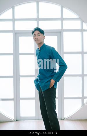 Un giovane e attraente asiatico posa con sicurezza in un ambiente studio, con un aspetto elegante e alla moda. Lui è in piedi rilassato di fronte a una grande finestra che indossa un Foto Stock