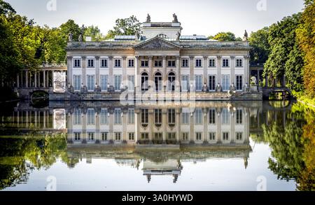 Un palazzo neoclassico con una facciata simmetrica si riflette in un tranquillo lago, circondato da lussureggiante vegetazione. L'edificio presenta colonne e decorazioni Foto Stock