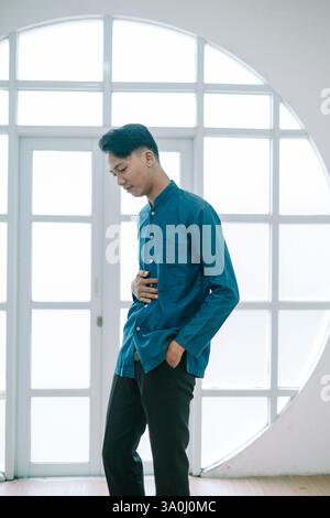 Un giovane uomo si staglia con cura in interni, indossando una camicia casual blu e pantaloni neri. Trasuda uno stile moderno, la sua posa contemplativa cattura Foto Stock