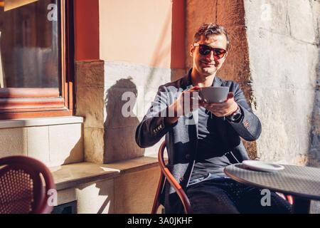 Ritratto di un uomo felice ed elegante che si rilassa nel caffè all'aperto bevendo un caffè. Il ragazzo guarda la macchina fotografica che offre bevande per strada Foto Stock
