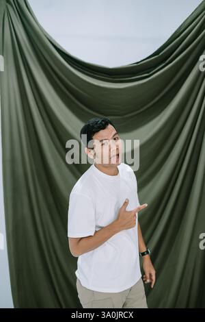 Un giovane asiatico si mette scherzosamente la lingua e punta a lato. Girato in studio con sfondo in tessuto verde drappeggiato. Indossa una t-S bianca Foto Stock