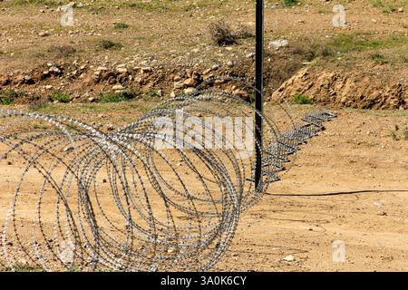 Le recinzioni di filo spinato a spirale si estendono lungo un terreno arido, mostrando costruzioni di confine in un ambiente naturale asciutto e polveroso con copertura limitata delle piante. Foto Stock