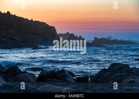 Le onde si infrangono contro la costa rocciosa mentre il sole tramonta su Matanzas in Cile, dipingendo il cielo con sfumature di arancione e viola. Foto Stock