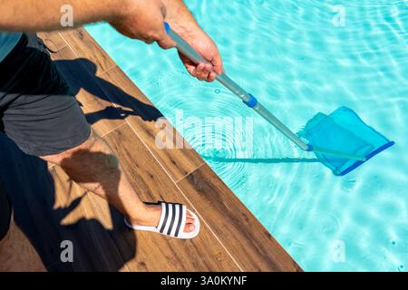 Uomo che pulisce la piscina turchese con rete skimmer, terrazza in legno e ambiente soleggiato all'aperto Foto Stock