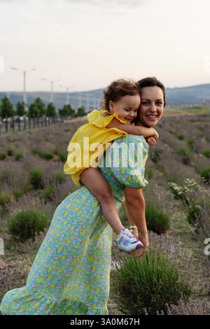 Madre e figlia si godono un gioioso picnic, corrono nel parco, giocano vicino al lago e si divertono in un campo di lavanda. Abiti luminosi, estate Foto Stock