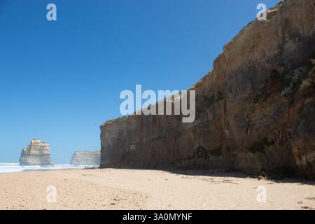 Spiaggia di Great Ocean Road: L'alta scogliera si innalza dalla spiaggia sabbiosa, le onde si infrangono sulla sabbia. In lontananza ci sono due pile di roccia. Cielo blu chiaro Foto Stock