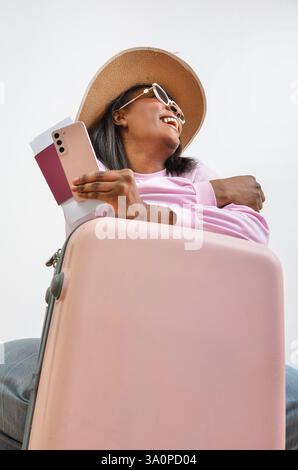 Il turista felice ha il passaporto e lo smartphone appoggiato alla valigia sorridente e rivolto verso l'alto Foto Stock