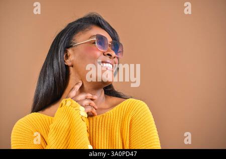Ritratto in studio di una allegra donna afroamericana che indossa occhiali da sole e un maglione giallo, toccando delicatamente il collo e guardando via con un sorriso Foto Stock