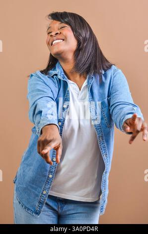 Donna felice che indossa una camicia in denim che punta le dita mentre sorride e guarda verso l'alto su sfondo marrone Foto Stock
