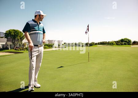 Uno sportivo si appoggia su un golf club su un putting green, osservando con attenzione il flagstick. Il cielo limpido e lo splendido paesaggio catturano l'essenza di Foto Stock