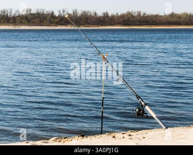 Canna da pesca con mulinello sulla riva sabbiosa di un ampio fiume. Foto Stock