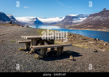 Tavolo da picnic in legno sulla riva di Isafjordur con montagne innevate e baia cristallina sullo sfondo, l'Islanda. Foto Stock