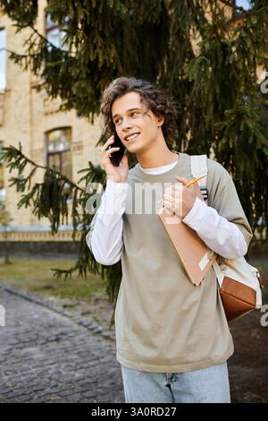 Lo studente elegante parla con piacere al telefono, portando con sé un notebook e una matita, per godersi il momento. Foto Stock