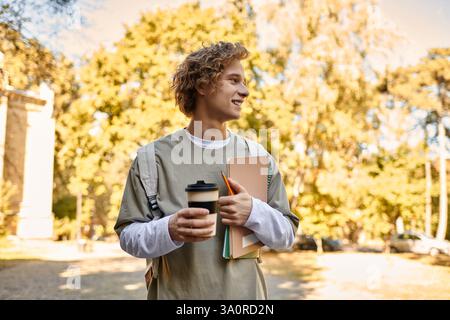 Il giovane sorride brillantemente mentre tiene in mano una tazza di caffè e libri, circondato da foglie autunnali. Foto Stock