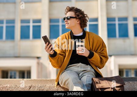 Un giovane uomo elegante si siede su una sporgenza, sorseggiando un caffè e sorridendo mentre scorre sul telefono. Foto Stock