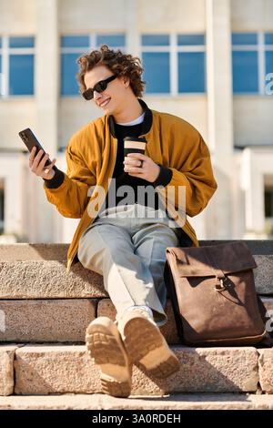 Un giovane alla moda siede su gradini di pietra, sorseggiando un caffè e sorridendo al telefono in piena luce del giorno. Foto Stock