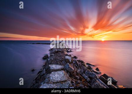 Un vivace tramonto illumina il cielo di Råbäck in Svezia, diffondendo tonalità calde sulle acque tranquille. Il molo roccioso si protende verso il mare, invitante Foto Stock