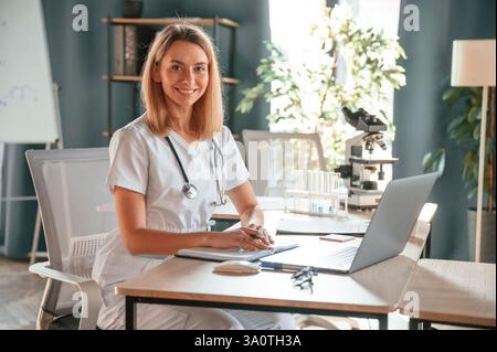 Umore sorridente e allegro. La giovane dottoressa in cappotto è seduta in clinica e sta lavorando. Foto Stock