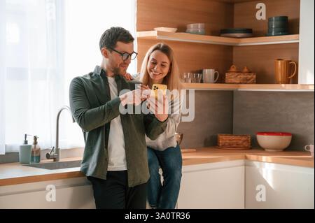 Uomo che mostra dei media a una donna al telefono. La giovane coppia è in cucina insieme. Foto Stock