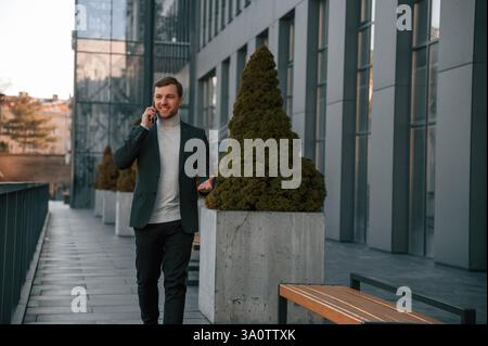 Vista frontale. Tramite smartphone. Un bell'uomo in abiti formali e' all'aperto vicino all'edificio degli affari. Foto Stock