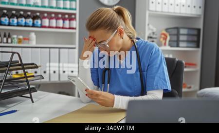 L'infermiera che indossa scrub blu e lo stetoscopio controlla il telefono che sembra stressato nell'interno organizzato di una clinica. Foto Stock