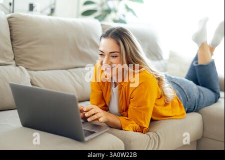 Una donna caucasica felice è comodamente sdraiata su un divano nel soggiorno, utilizza il suo laptop e sorride mentre invia messaggi online con gli amici, mentre l'umore rilassato e positivo si mostra mentre si diverte a casa Foto Stock