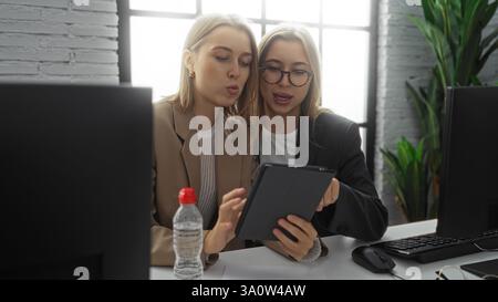 Le donne che collaborano in un ufficio, recensiscono il lavoro su un tablet, indossano abiti da lavoro, circondate da computer e piante, al chiuso, appaiono pubbliche relazioni Foto Stock